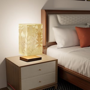 May include: A bedside table lamp with a square, cream-colored lampshade featuring a floral pattern. The lamp is on a wooden base and is sitting on a wooden nightstand with two drawers.