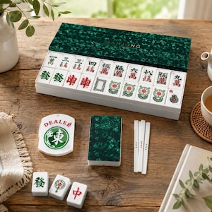 K&ouml;nnte beinhalten: Ein komplettes Mahjong-Set mit einer gr&uuml;n strukturierten Box und wei&szlig;en Spielsteinen mit roten und gr&uuml;nen Markierungen. Das Set enth&auml;lt einen "DEALER"-Marker, W&uuml;rfel und ein kleines gr&uuml;nes Buch. Eine Teekanne, eine Teetasse und ein Buch mit dem Titel "The Kinfolk Garden" sind ebenfalls vorhanden.