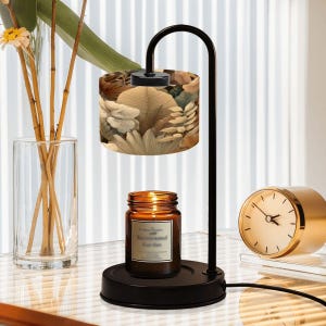May include: A black metal candle warmer with a floral patterned lampshade. A lit candle in a glass jar sits on the base. A gold-colored clock is on the right. The candle jar label reads "Among scents, Recreated Garden."