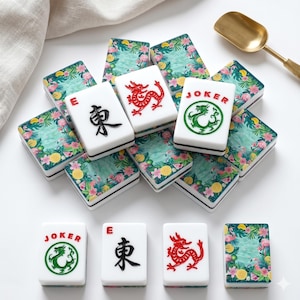 May include: A collection of white Mahjong tiles with black, red, and green symbols. The tiles are decorated with a tropical floral design. A gold spoon rests in the background.