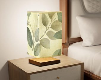 May include: A bedside table lamp with a square, cream-colored lampshade featuring a green leaf pattern. The lamp is on a wooden base and sits on a wooden nightstand with two drawers.