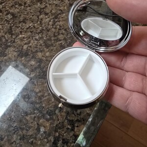 Puede incluir: Un peque&ntilde;o pastillero redondo y plateado con tapa con bisagras y espejo. El interior tiene tres compartimentos blancos. El estuche est&aacute; abierto, revelando los compartimentos. El estuche se sostiene en la mano de una persona.