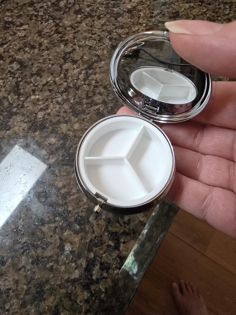 Puede incluir: Un peque&ntilde;o pastillero redondo y plateado con tapa con bisagras y espejo. El interior tiene tres compartimentos blancos. El estuche est&aacute; abierto, revelando los compartimentos. El estuche se sostiene en la mano de una persona.
