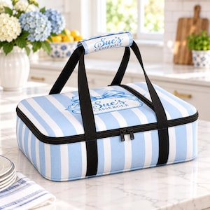 May include: A rectangular insulated casserole carrier featuring a light blue and white vertical striped pattern. The top lid displays a blue bow graphic and the text Sue's Casserole. It has black carrying straps and a padded handle with the same text.