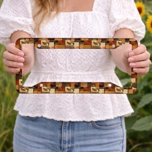 May include: License plate frame with a repeating pattern of squares in shades of brown, tan, and green, featuring leaves and geometric shapes. The frame has a rectangular shape with rounded corners and holes for mounting.