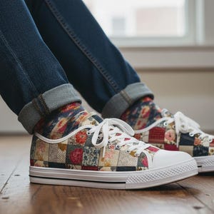 May include: Low-top trainers with a patchwork design featuring floral and geometric patterns in red, pink, blue, and cream colours. The shoes have white laces, a white rubber toe cap, and a white sole. The person is wearing blue jeans.