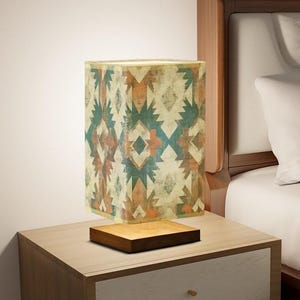 May include: A square-shaped table lamp with a geometric patterned lampshade in shades of brown, green, and white. The lamp is sitting on a wooden base and is on a wooden nightstand.