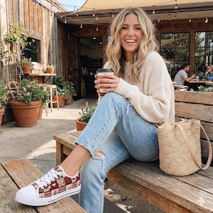 May include: White canvas sneakers with a red and white patchwork design. The sneakers have white laces and a white rubber sole. The shoes are worn by a person sitting on a wooden bench.