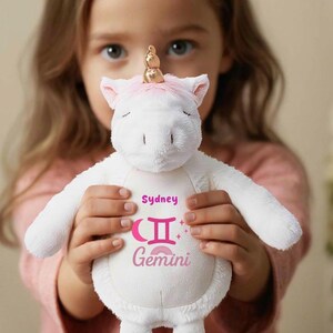 May include: A white plush unicorn toy with pink accents, including the ears and hooves. The unicorn has a golden horn and the name "Sydney" and the word "Gemini" with the Gemini symbol printed on its belly.
