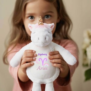 May include: A white plush unicorn toy with pink accents on the ears and hooves. The unicorn has a gold horn and the name "Sydney" and the word "Aries" are printed on the body. The toy is held by a person.