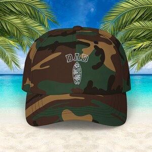 May include: Camouflage baseball cap with the word "DAD" in white block letters above a white surfboard design. The cap features a curved brim and is set against a tropical beach scene.