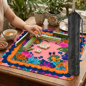 May include: A Mahjong game in progress on a pink floral mat with green and white tiles. The mat has a blue and orange border. A black carrying case is on the right. A person is playing the game.