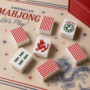 May include: A set of white Mahjong tiles featuring red striped patterns, a red dragon, a green dragon, and a Joker tile with a green dragon graphic. The tiles are arranged on a beige surface printed with the text American Mahjong Let's Play.
