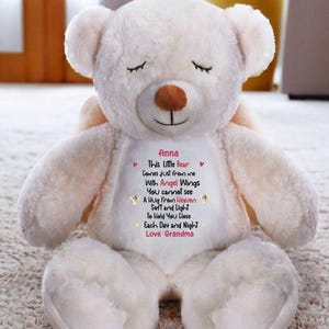May include: A cream-colored teddy bear with a brown nose and closed eyes. The bear has a white patch on its chest with the text "Anna This Little Bear Comes just from me With Angel Wings You cannot see A Hug from Heaven Soft and Light To Hold You Close Each Day and Night Love Grandma."