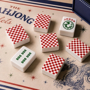 May include: Six white rectangular mahjong tiles with red and white checkered backs, one tile featuring a green dragon and the word JOKER, and another with green bamboo symbols. The tiles rest on a mat printed with THE MAHJONG and a dragon illustration.