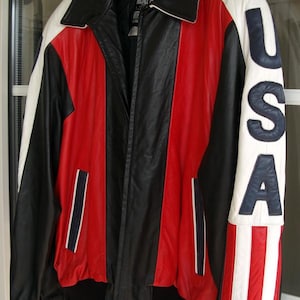 Könnte beinhalten: Eine schwarze, rote und weiße Lederjacke mit Reißverschluss. Die Jacke hat einen weißen Streifen am linken Ärmel mit dem Wort "USA" in blauen Buchstaben. Die Jacke hat einen schwarzen Kragen und einen schwarzen Reißverschluss.