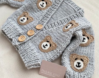 May include: Gray knitted baby cardigan with brown bear appliques. The cardigan has a button closure and is made with a soft, cozy yarn.