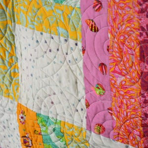 May include: A patchwork quilt featuring vertical strips of fabric in vibrant colours including yellow, pink, orange, and teal. The fabric displays various patterns such as small flowers, abstract shapes, and shells. The quilt has a continuous curved stitching pattern.