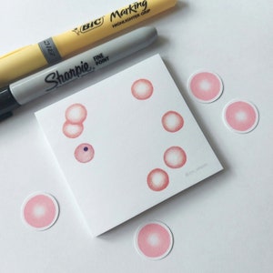 May include: A white square notepad with pink dot illustrations and several pink circle stickers. A yellow highlighter and a grey Sharpie marker are positioned above the notepad.