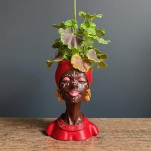 May include: A dark brown decorative planter in the shape of a woman's head, with a red head wrap and earrings. The planter holds a green and purple plant. The face has white markings and closed eyes. The bust is red. The planter is suitable for indoor use.