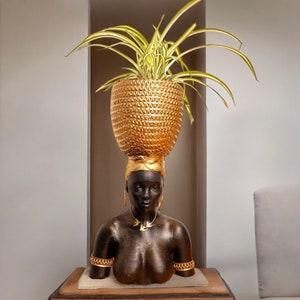 May include: A gold planter with a textured surface sits atop a black bust with gold accents. The planter is filled with a green plant with long, thin leaves.