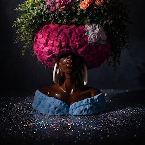 May include: A decorative bust with a dark brown face, large white earrings, and a blue off-the-shoulder top. The bust is topped with a large, pink, textured headpiece and a vibrant arrangement of flowers in shades of pink, orange, and green.