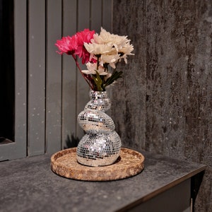 Könnte beinhalten: Eine silberne, vielseitige Vase mit einem runden, bauchigen Boden und einem zylindrischen Hals hält einen Strauß aus rosa und weißen Blumen. Die Vase steht auf einem braunen, hölzernen Untersetzer.