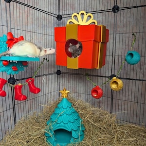 Könnte beinhalten: Ein Rattenkäfig, der für Weihnachten dekoriert ist, mit einem türkisfarbenen Weihnachtsbaumhaus, einem roten Geschenkboxhaus und hängenden Ornamenten. Eine weiße und graue Ratte befindet sich auf einem türkisfarbenen Regal mit einer roten Schleife.