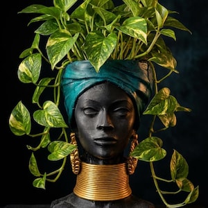 Puede incluir: Un macetero con forma de busto negro que representa a una persona con un turbante azul verdoso y un collarín dorado ancho. La cabeza sirve como recipiente para una planta potos colgante con hojas acorazonadas de color verde y amarillo.