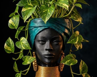 May include: A black bust planter featuring a person wearing a teal headwrap and a wide gold neck ring. The head serves as a container for a trailing pothos plant with variegated green and yellow heart-shaped leaves that drape over the sides.
