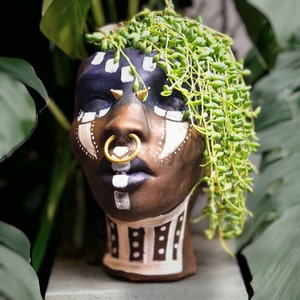 Puede incluir: Un macetero de cerámica marrón con forma de cabeza humana con patrones geométricos blancos y negros pintados en la cara. El macetero tiene un aro de nariz dorado y está decorado con una planta suculenta verde.
