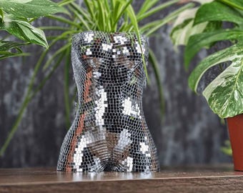 May include: A silver mirrored planter shaped like a human torso, with a green plant growing out of the top.