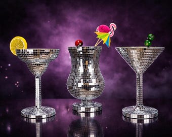 May include: Three cocktail glasses covered in mirrored tiles, each with a different shape and garnish. One has a lemon slice, another a cherry, and the third has two green olives. The background is a dark purple.