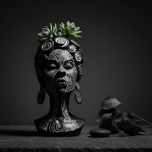 May include: A black and white decorative head planter with detailed spiral and geometric patterns. The planter holds small green succulents. The head has earrings and a detailed face. A dark background and leaves and stones complete the scene.