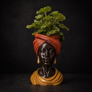 May include: A decorative planter shaped like a woman's head, featuring a turban and earrings. The planter is filled with green leafy plants. The head is dark brown with a yellow and orange scarf and earrings.