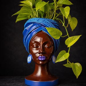 May include: A decorative brown head-shaped planter with a blue turban and matching earrings. The lips are painted purple. The planter holds green leafy plants. The base and the turban have a blue geometric design.