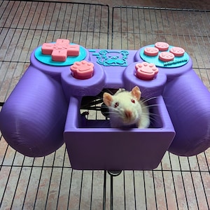 Puede incluir: Un hábitat morado para mascotas con forma de mando de videojuegos. El mando tiene botones rosas y turquesas, con una pequeña rata blanca dentro del compartimento central. El hábitat está sobre una rejilla metálica.