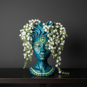 Puede incluir: Una escultura decorativa de cabeza de color turquesa con detalles amarillos y un arreglo floral blanco. La escultura presenta un rostro estilizado con ojos cerrados y patrones intrincados. Las flores caen en cascada.