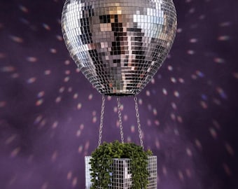 May include: A mirrored disco ball planter with a square, mirrored planter box suspended below. The planter box is filled with green, trailing plants. The background is a dark purple with light reflections.