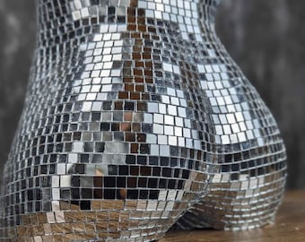 以下が含まれることがあります： 小さな鏡タイルで作られた銀色のディスコボール型の彫刻。彫刻は人間の胴体のような形をしており、木製の表面の上に置かれています。