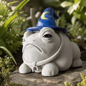 May include: A light grey concrete frog figurine wearing a blue wizard hat. The hat features a yellow crescent moon and small yellow stars. The frog has a smooth, rounded body, a wide mouth, and dark eyes, with a thin cord tied around its neck.