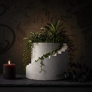 Op de afbeelding: Een cilindrische betonnen plantenbak met een spiraalvormig trapontwerp, gevuld met groen. Een brandende bordeauxrode kaars staat links. De achtergrond is een donkere, getextureerde muur. De plantenbak heeft een diameter van ongeveer 30 cm.