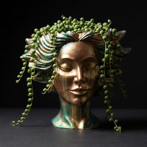 May include: A decorative head-shaped planter with closed eyes, painted in gold and teal. The planter is filled with a cascading string of pearls plant, creating a unique and artistic display. The planter is a striking piece of home decor.