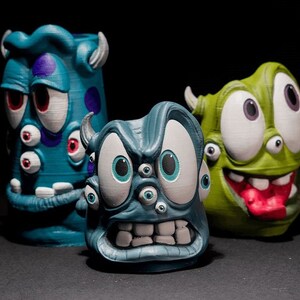 May include: Three 3D printed figurines of cartoon monsters with multiple eyes. The first monster is blue with purple spots and red eyes. The second monster is blue with white eyes and a toothy grin. The third monster is green with white eyes and a red tongue sticking out.