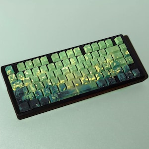 May include: A mechanical keyboard featuring a black frame and custom keycaps with a gradient color scheme transitioning from light sage green at the top to dark forest green and black at the bottom, accented with yellow paint splatter details.