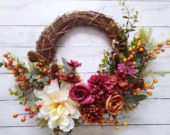 May include: A brown twig wreath decorated with autumn-coloured flowers, including white peonies, pink roses, and burgundy dahlias. The wreath is accented with sprigs of greenery and red and orange berries.