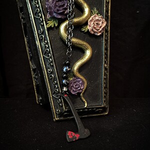 May include: A black coffin-shaped box with gold trim and a gold snake with purple and pink flowers. A black chain with black beads and a black axe with red paint hangs from the box.