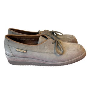 May include: A pair of gray suede Mephisto shoes with laces. The shoes have a rounded toe and a brown trim. The Mephisto logo is visible on the side of the shoe. The shoes are a size 6.5.