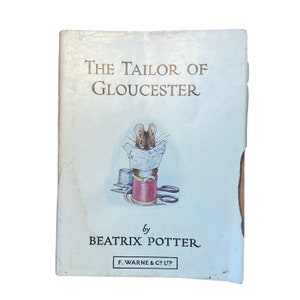 Puede incluir: Una portada de libro vintage para "El sastre de Gloucester" de Beatrix Potter. La portada presenta un fondo azul claro con un ratón marrón sentado en un carrete de hilo rojo, leyendo un periódico.