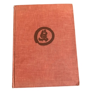 May include: A vintage book with a textured, reddish-brown cover. A dark brown circular emblem with a Japanese character is centered on the front. The book's spine is visible on the left side, suggesting it is a hardcover book.